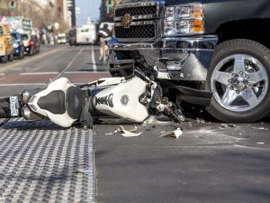 A motorcycle is on its side, underneath a large truck. Parts of motorcycle are spread out on the road.