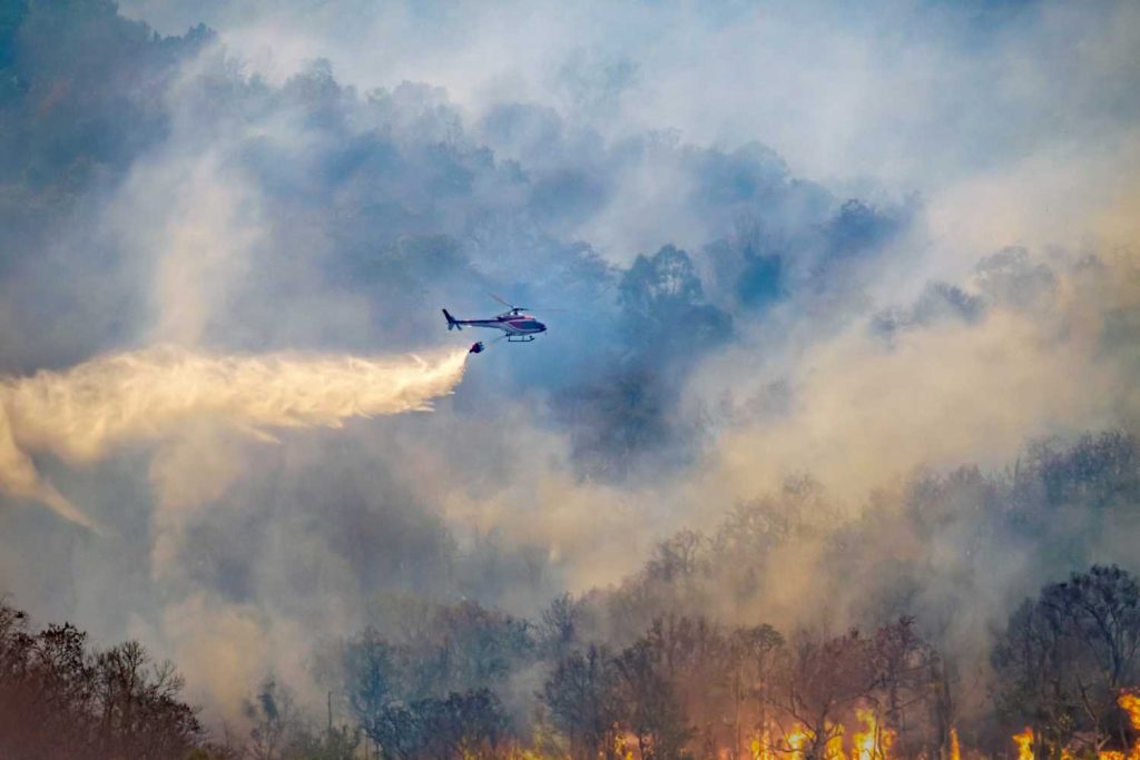 A fire helicopter flies over a forest fire dropping water.